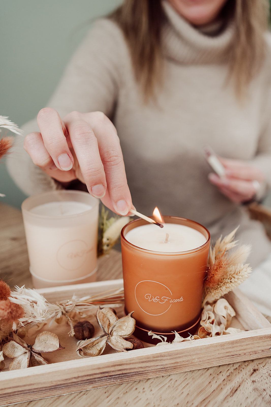 Hand steekt V&F Scents geurkaars aan in oranje glazen pot op houten dienblad